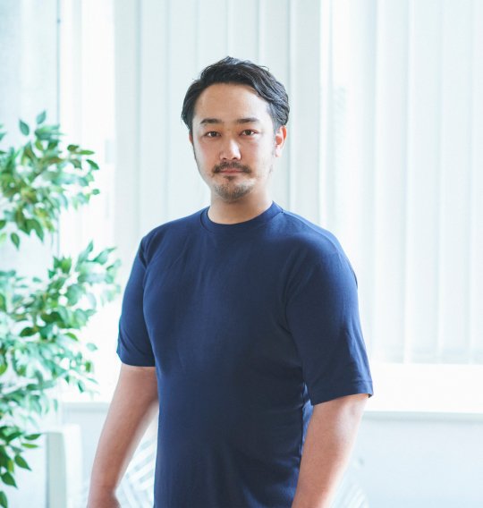 明るい室内で、ネイブーのTシャツを着た男性が落ち着いた表情でまっすぐ立っている。堂々とした佇まいが印象的で、背景には観葉植物とブラインドが見える。