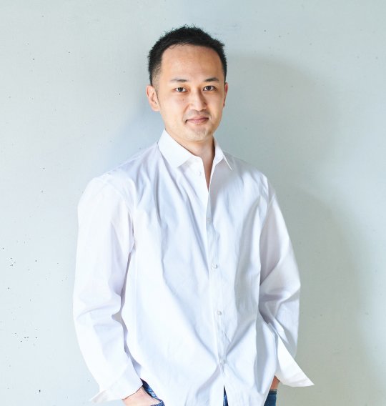 明るい背景の前で、白いシャツ姿の人物が穏やかな表情で立っている。