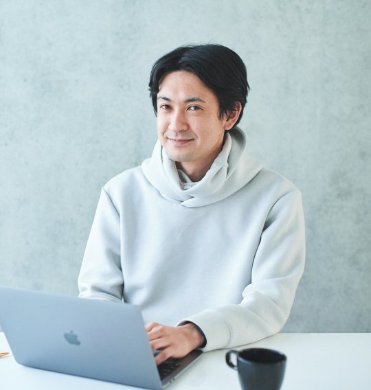 明るい室内で、薄いグレーのパーカーを着た人物がノートPCに手を添えてこちらを見ている。机の上にマグカップが置かれている。