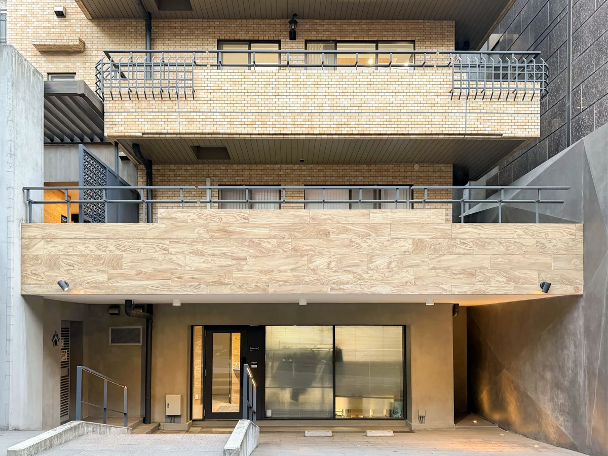 コンクリートの壁と石の階段が続き、その先に建物の明るいエントランスが見える。静かで落ち着いた雰囲気のアプローチが印象的な写真。