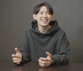 笑顔で座って話している人物の写真。ダークグレーのパーカーを着た男性の笑顔が印象的なシーン。